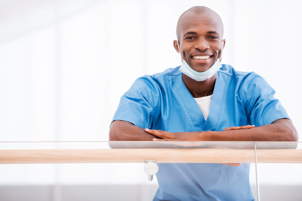 Cheerful doctor. Low angle view of confident young African doctor in blue uniform looking at camera and smiling while leaning at the handrail